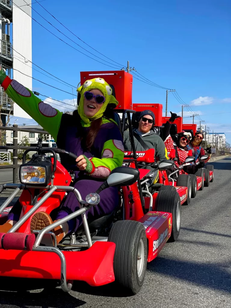 IMG_1486 - StreetKart Person in a bright frog hat and purple suit drives a red go-kart, waving at the camera; riders in costumes follow behind on a sunny street.