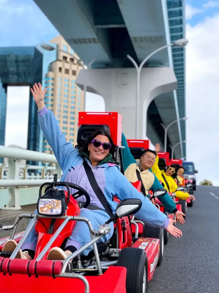 IMG_2914 - StreetKart Group of people riding red go-karts under a large elevated bridge, front rider in a blue jacket waving at the camera.