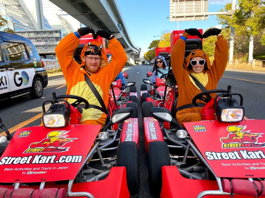 Two people in orange animal onesies in red go-karts under a city overpass, making heart shapes with their arms toward the camera; Street Kart branding visible on the carts' fronts.